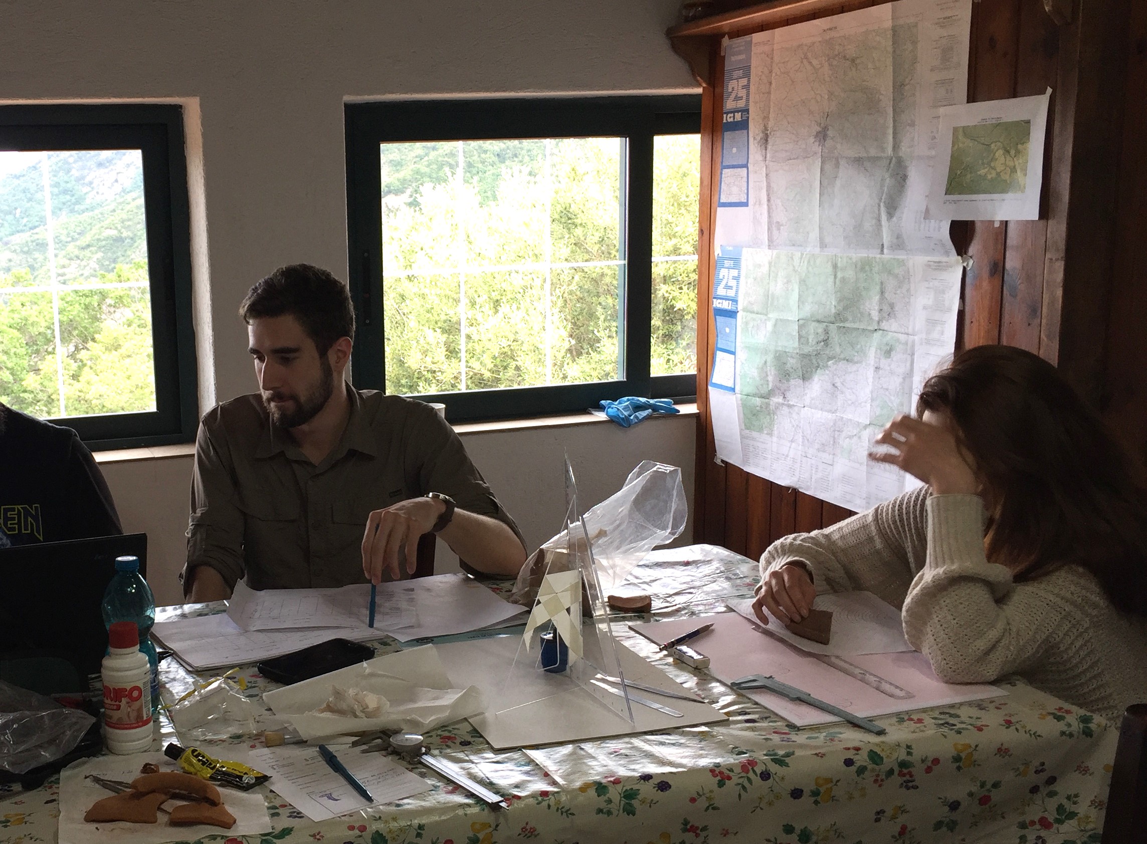 a man and a woman are sitting at a table working on archaeological material with drawing implements and a computer