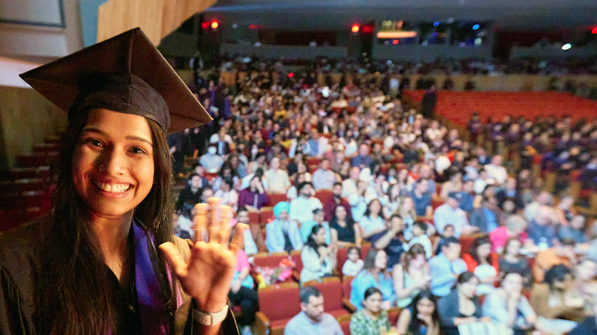 A female student in cap and gown waves and smiles at the camera as she crosses the stage.